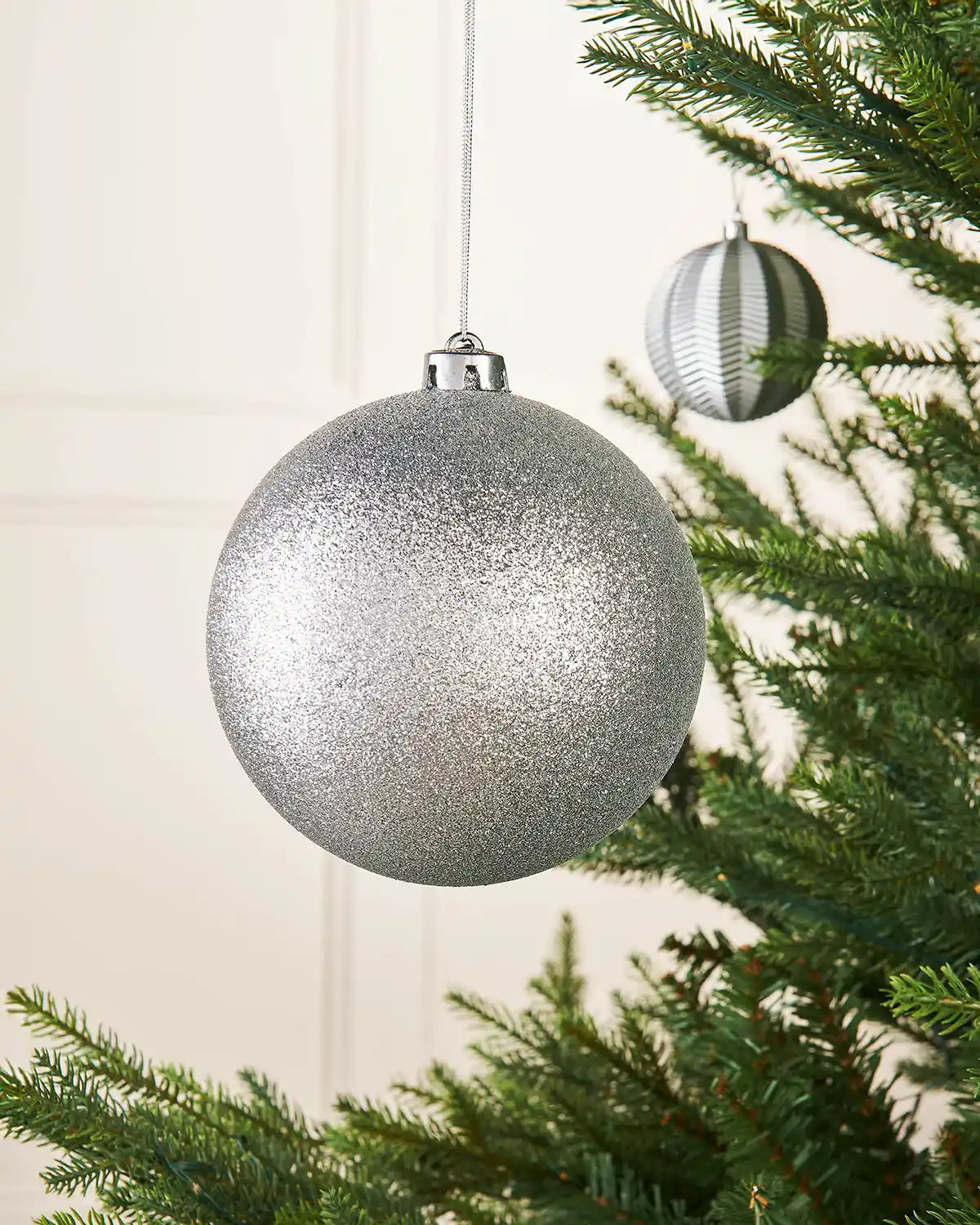 Silver oversized 15cm glitter bauble on a Christmas tree branch with a smaller silver bauble in background