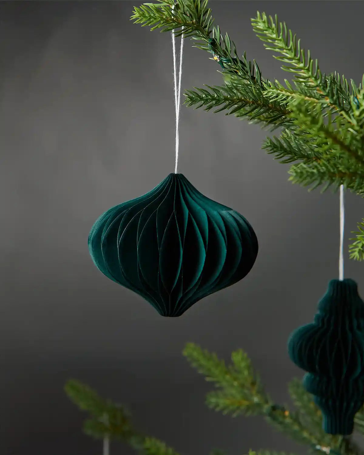 A single 10cm sage green honeycomb paper tree decoration hanging on Christmas tree in front of grey wall background