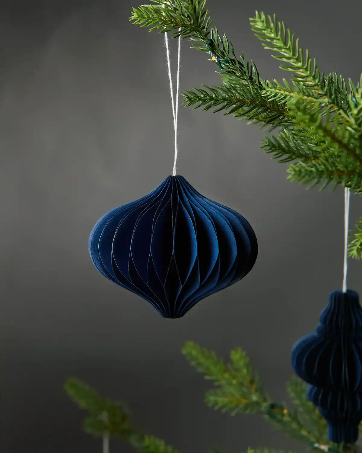 Image of a single bulb-shaped 10cm navy blue paper tree decoration hanging on a Christmas tree in front of grey wall background