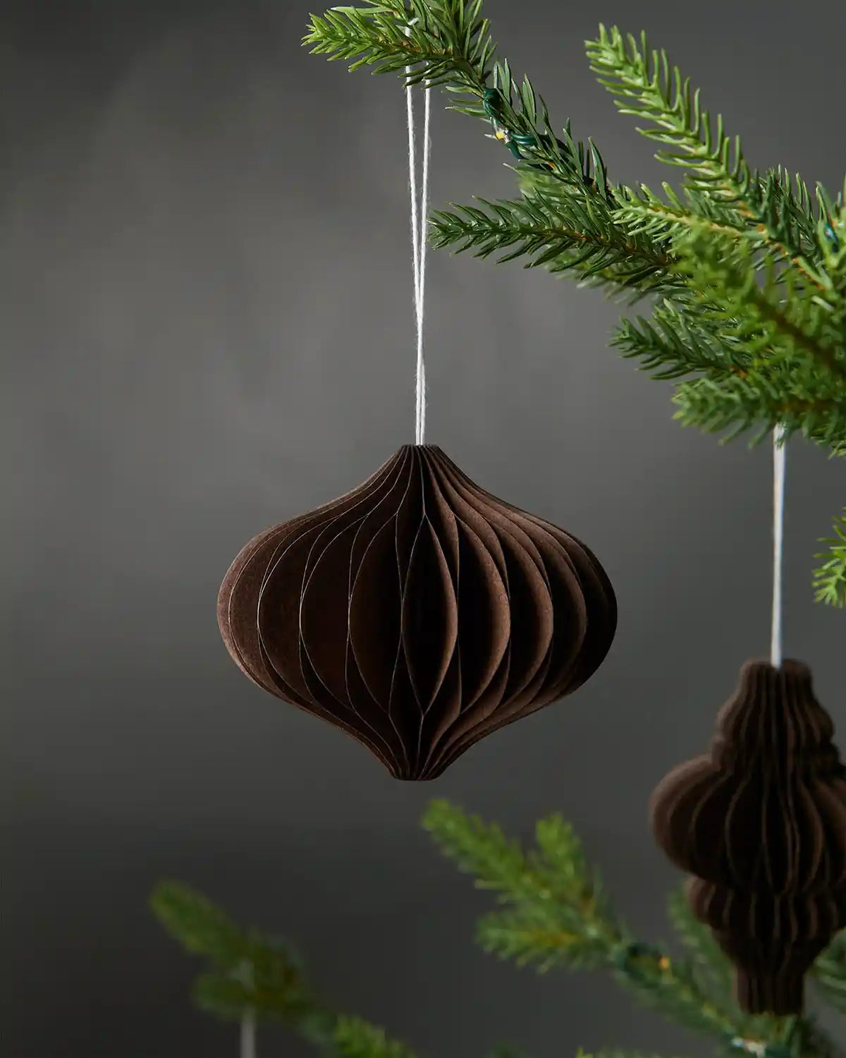 Image of a single bulb-shaped espresso brown paper honeycomb tree decoration hanging on Christmas tree in front of grey wall background