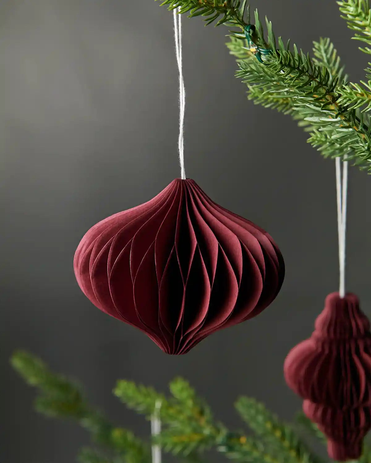 Close-up of single bulb-shaped burgundy red paper tree decoration hanging on tree in front of grey wall background