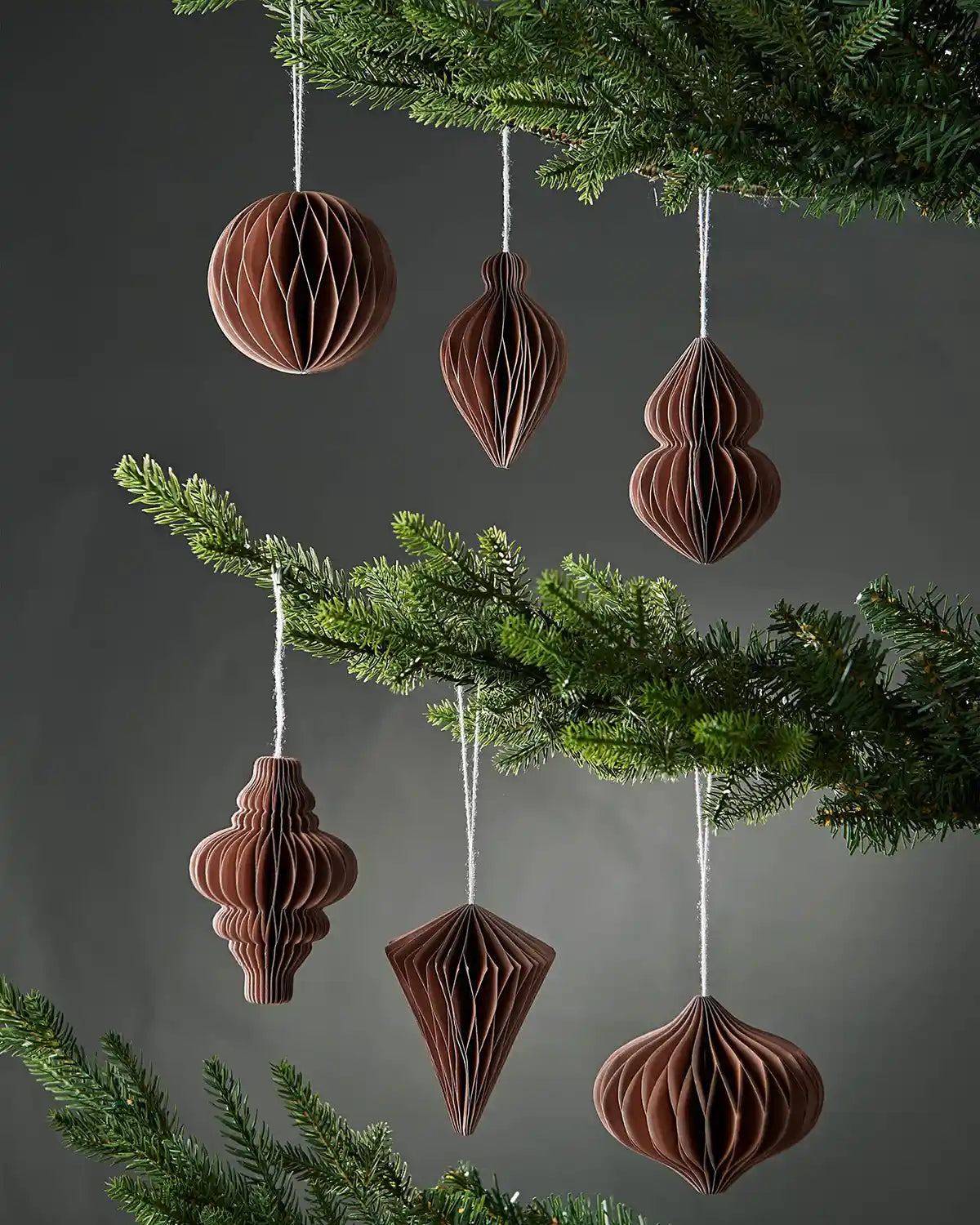 Six 10am mocha brown paper Christmas decorations hanging on Christmas tree branches in front of a grey wall background