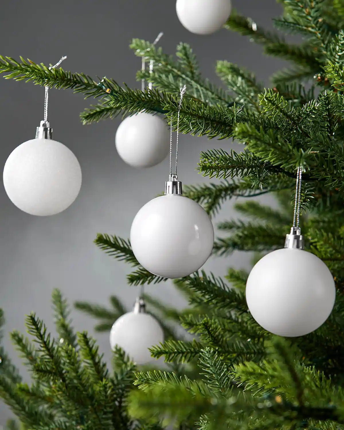 Six White Christmas baubles hanging on a Christmas tree branch against a grey background