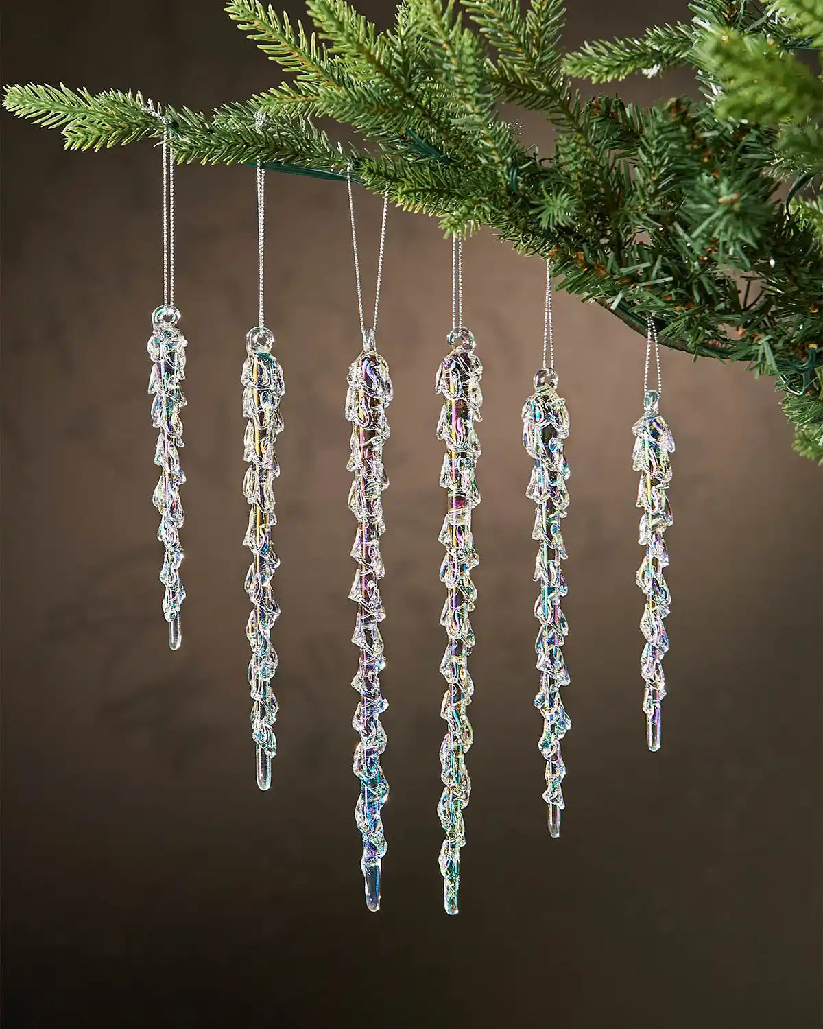 Six iridescent crystal icicle ornaments hanging from a Christmas tree branch against a brown background  