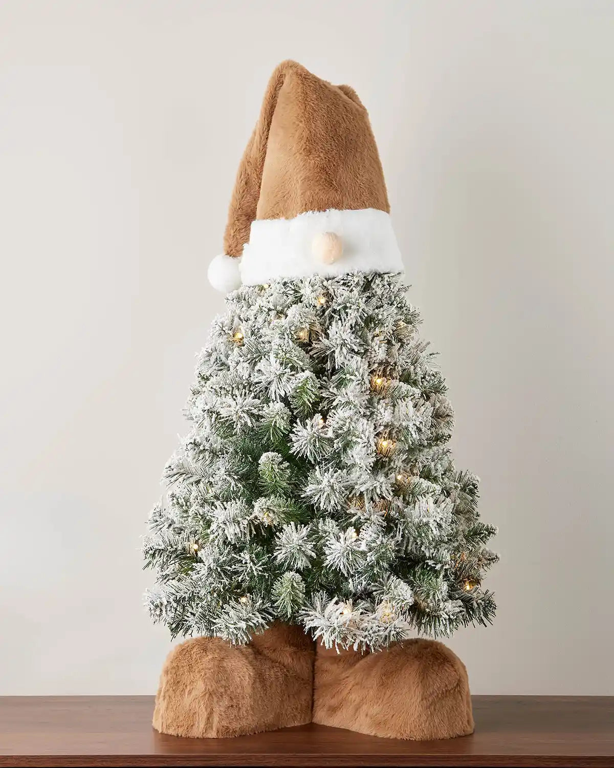 Small gonk Christmas tree with Brown Santa hat and brown boots against a white wall on a wooden table