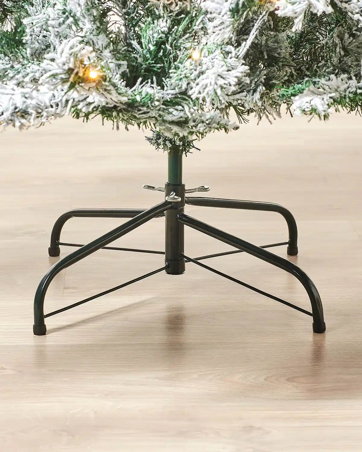 Snow flocked 7ft artificial Christmas tree with black stand on a wooden floor.