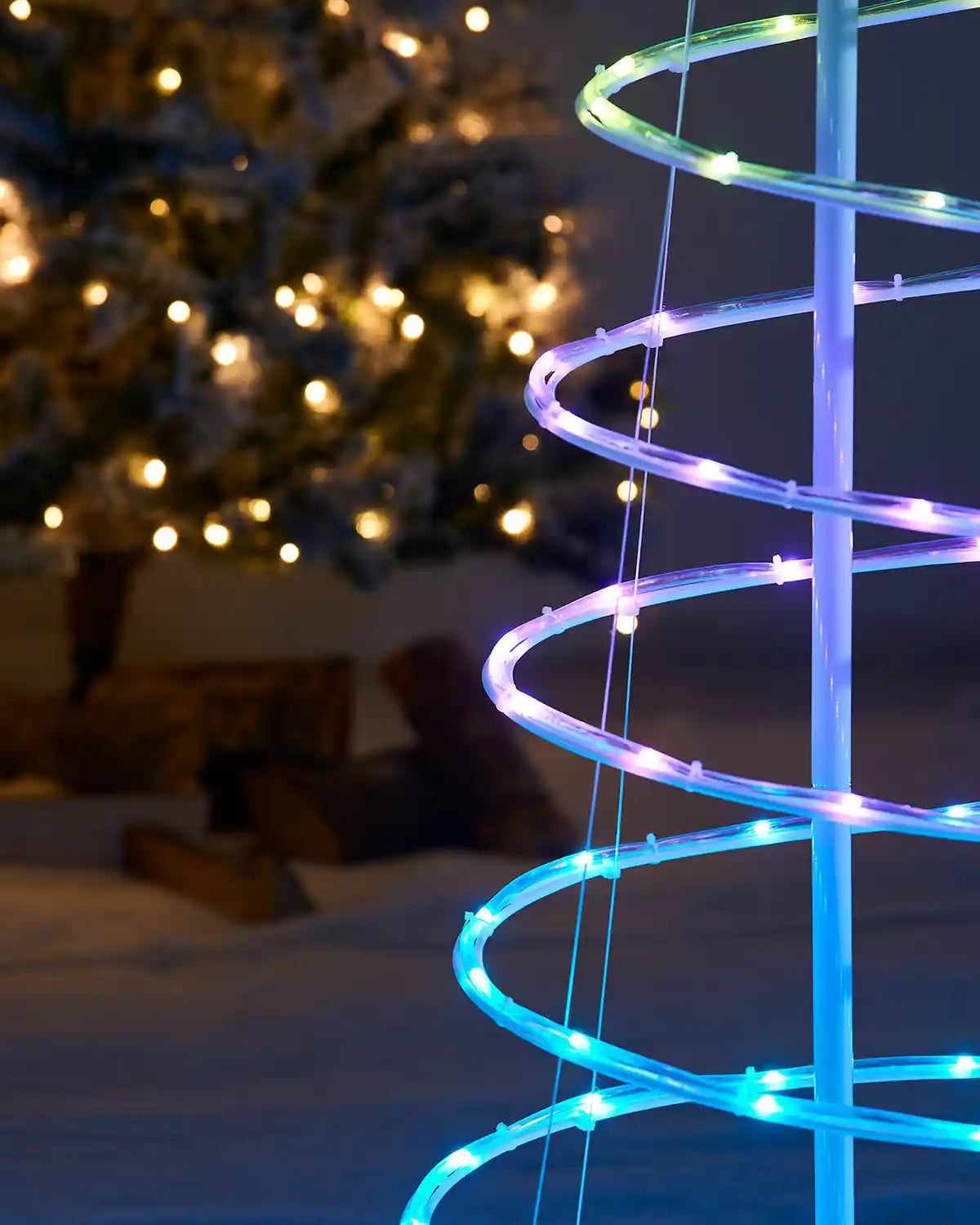 Spiral green, purple and blue LED light decoration in front of a Christmas tree with lights.