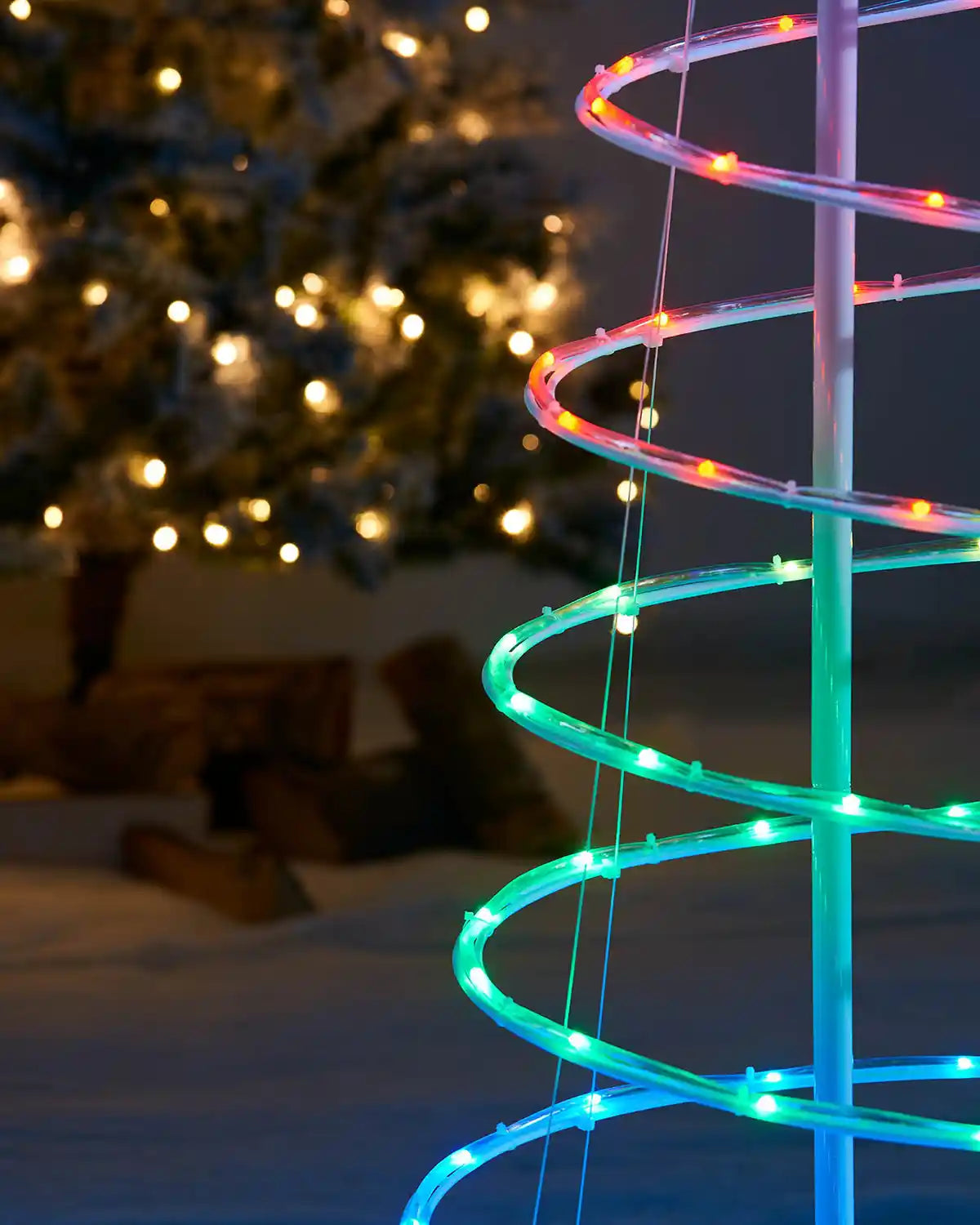 Spiral tree red, green and blue LED light decoration in front of a Christmas tree with lights.