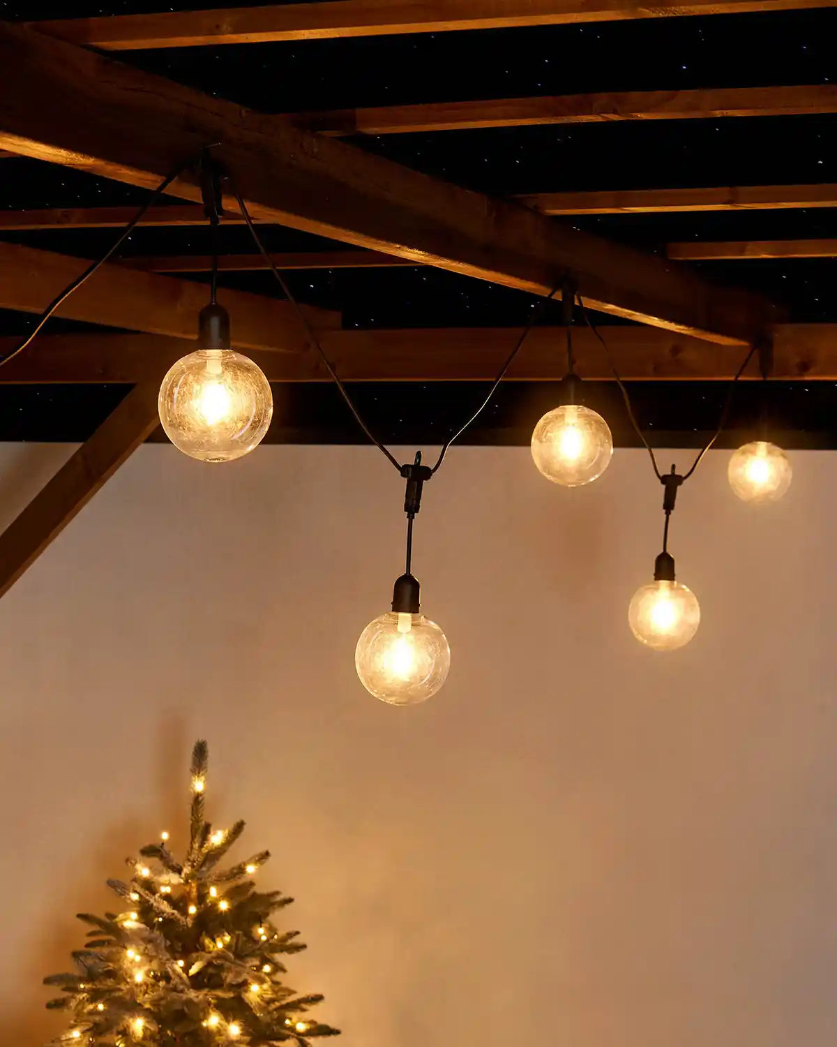 String of light bulbs hanging from a wooden ceiling with a Christmas tree in the background.