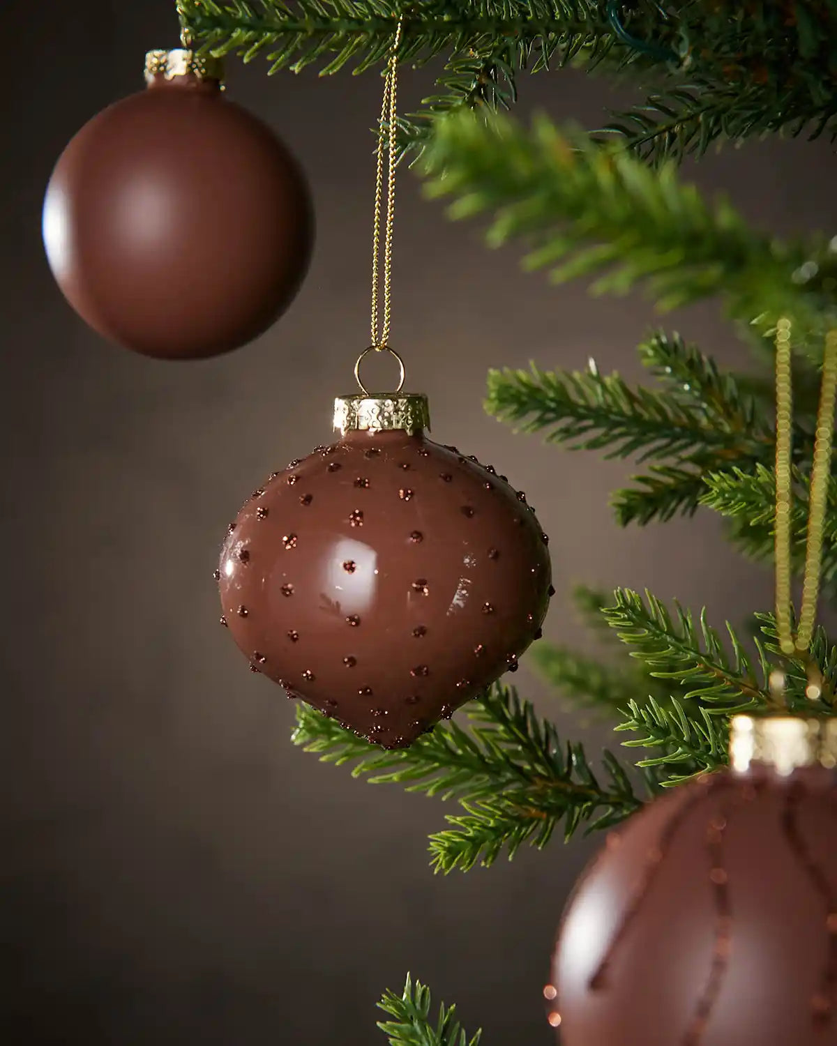 Three Brown textured Christmas baubles hanging on a tree with a dark background