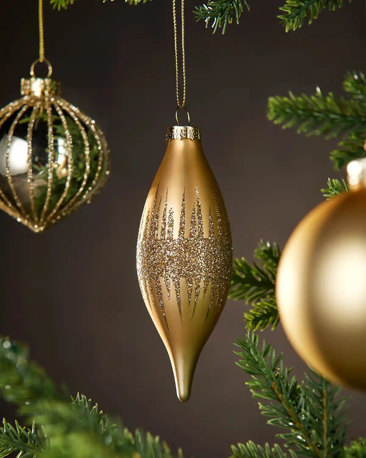 Three Gold Christmas baubles in various shapes and textures hanging on a tree with a dark background