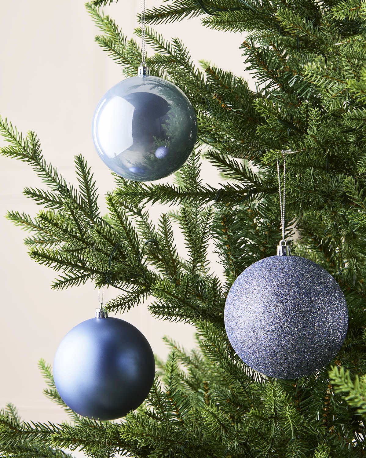 Three Ice blue Christmas tree ornaments hanging on a green evergreen tree against a light beige background.