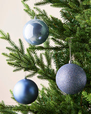 Three Ice blue Christmas tree ornaments hanging on a green evergreen tree against a light beige background.