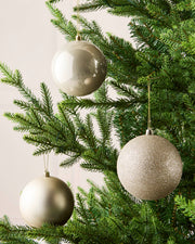 Three Latte holiday tree ornaments in champagne, gold, and silver tones hanging from the green branches of a Christmas tree.