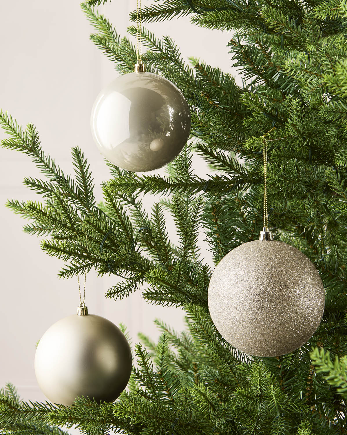 Three Latte holiday tree ornaments in champagne, gold, and silver tones hanging from the green branches of a Christmas tree.