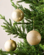 Three Latte holiday tree ornaments in champagne, gold, and silver tones hanging from the green branches of a Christmas tree.
