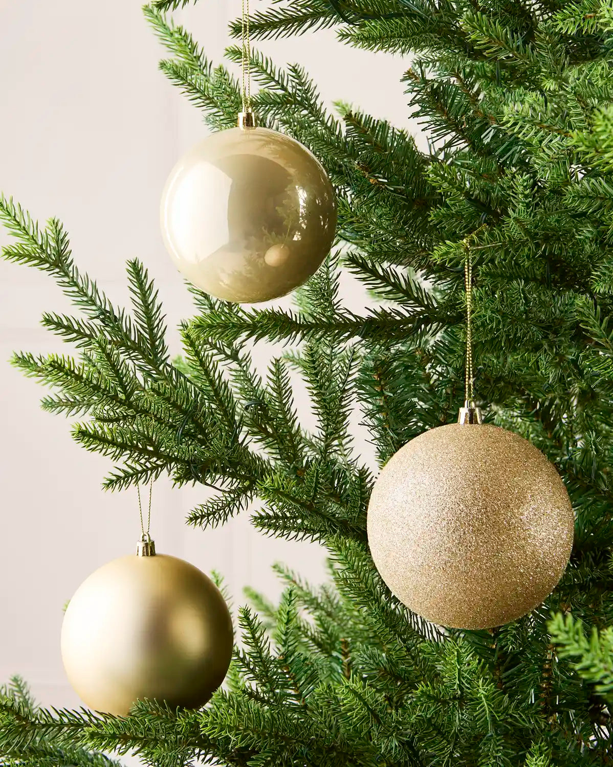 Three Latte holiday tree ornaments in champagne, gold, and silver tones hanging from the green branches of a Christmas tree.
