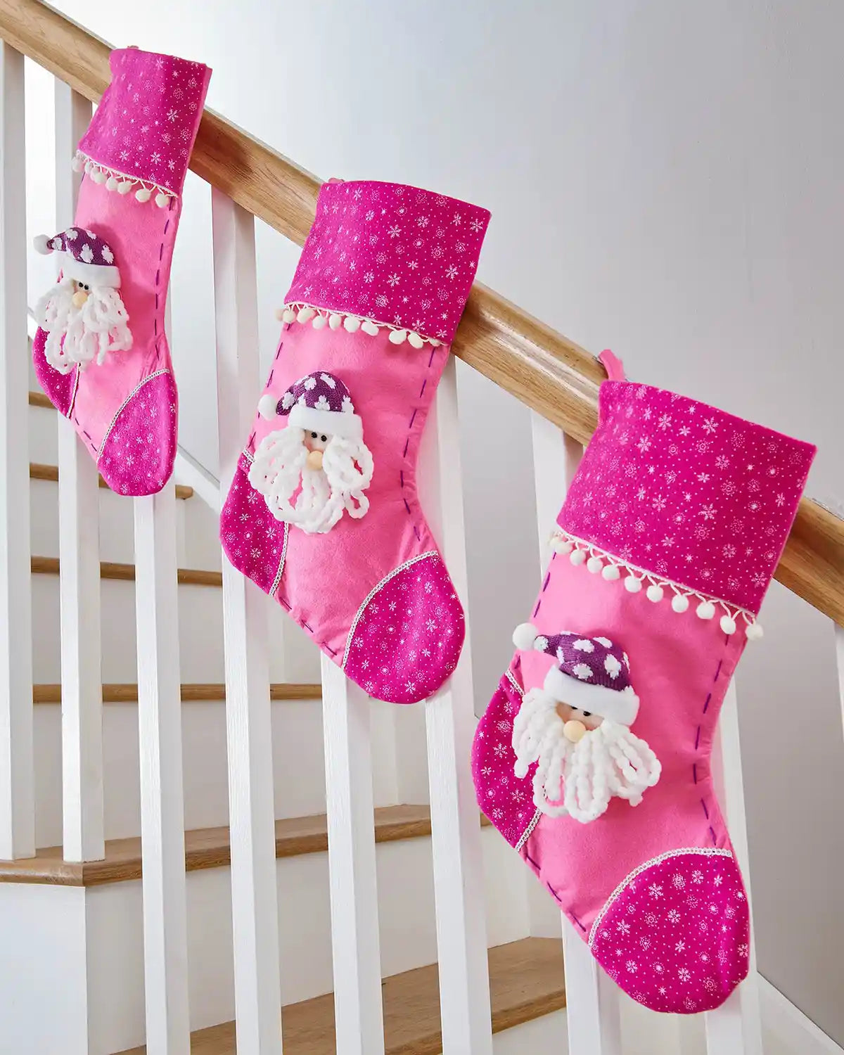 Three Pink Christmas stockings with 3D Santa Claus face and snowflake pattern on a wooden staircase banister. 
