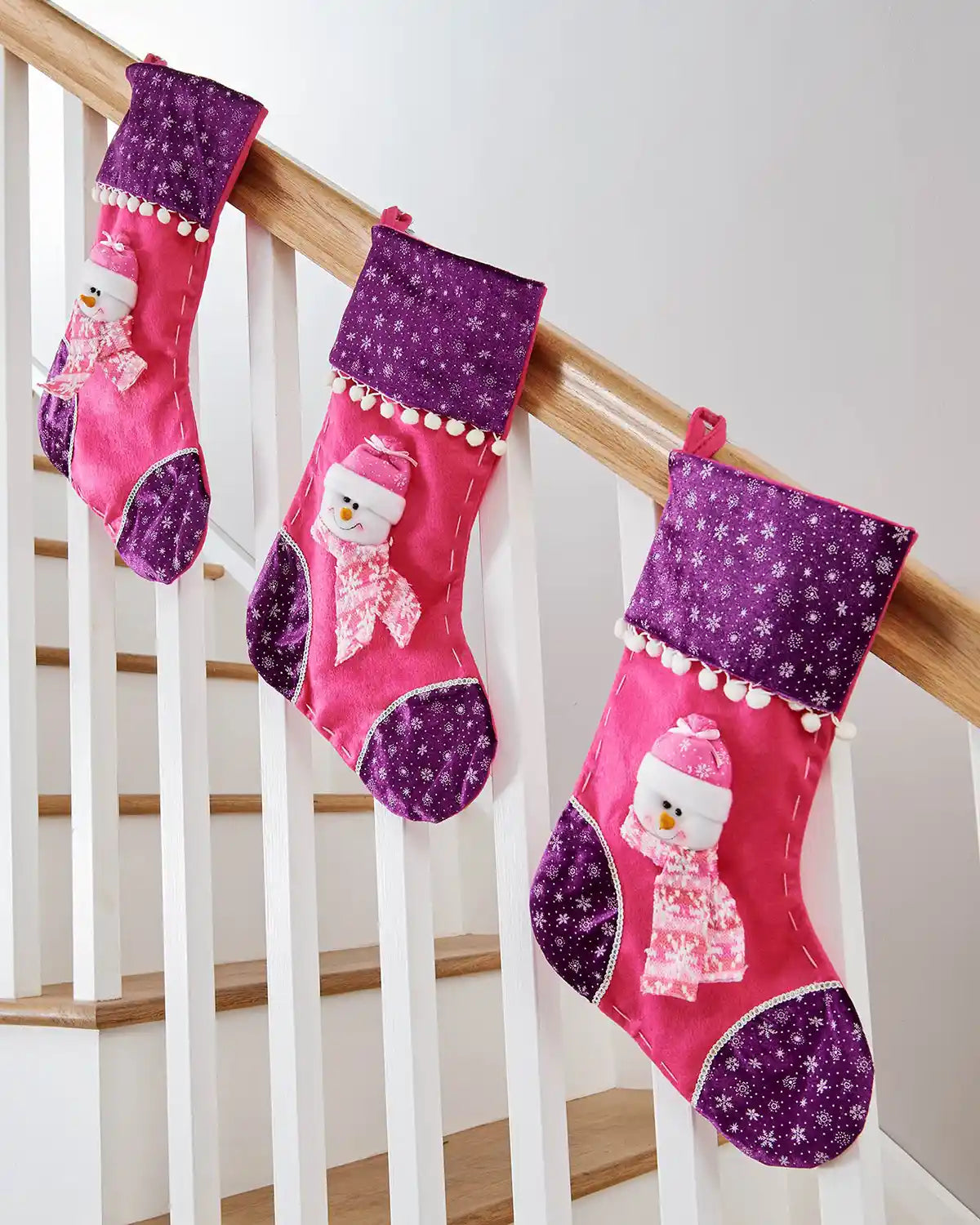 Three Pink and Purple Christmas stocking with 3D Snowman with Pink scarf and snowflake pattern with pom poms on a wooden staircase banister 
