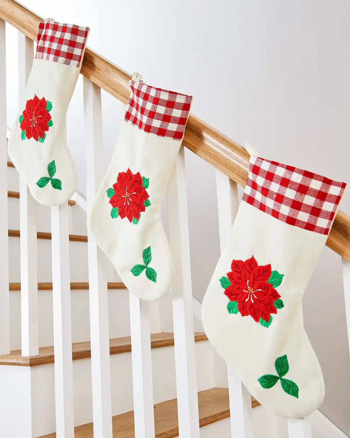Three Christmas stockings with red Poinsettia flowers and Tartan trim hanging on a wooden staircase banister. 