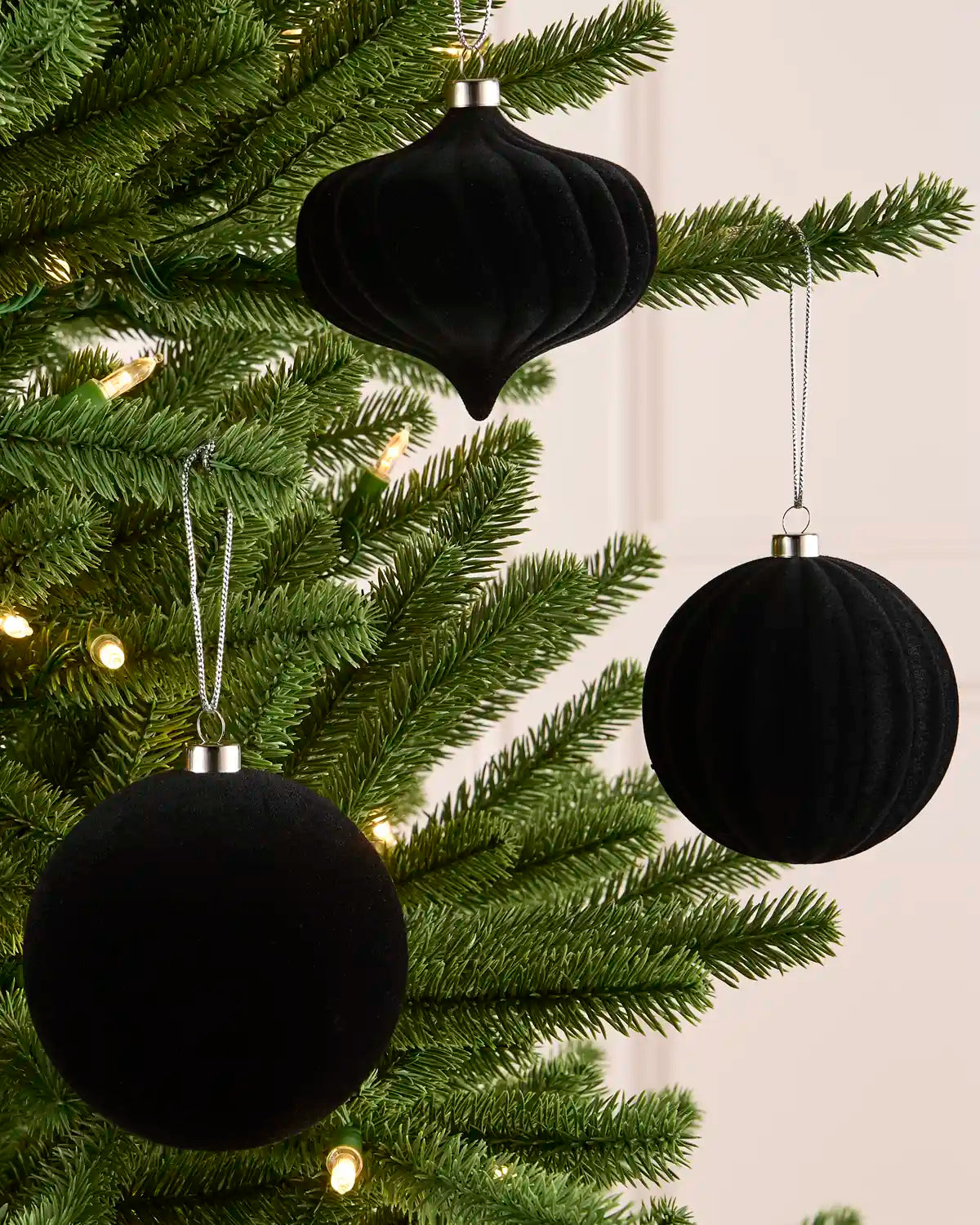 Three black velvet Christmas tree ornaments, including two round baubles and one teardrop-shaped ornament, hanging on a green pine tree with warm string lights.