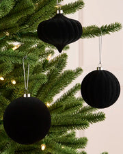 Three black velvet Christmas tree ornaments, including two round baubles and one teardrop-shaped ornament, hanging on a green pine tree with warm string lights.