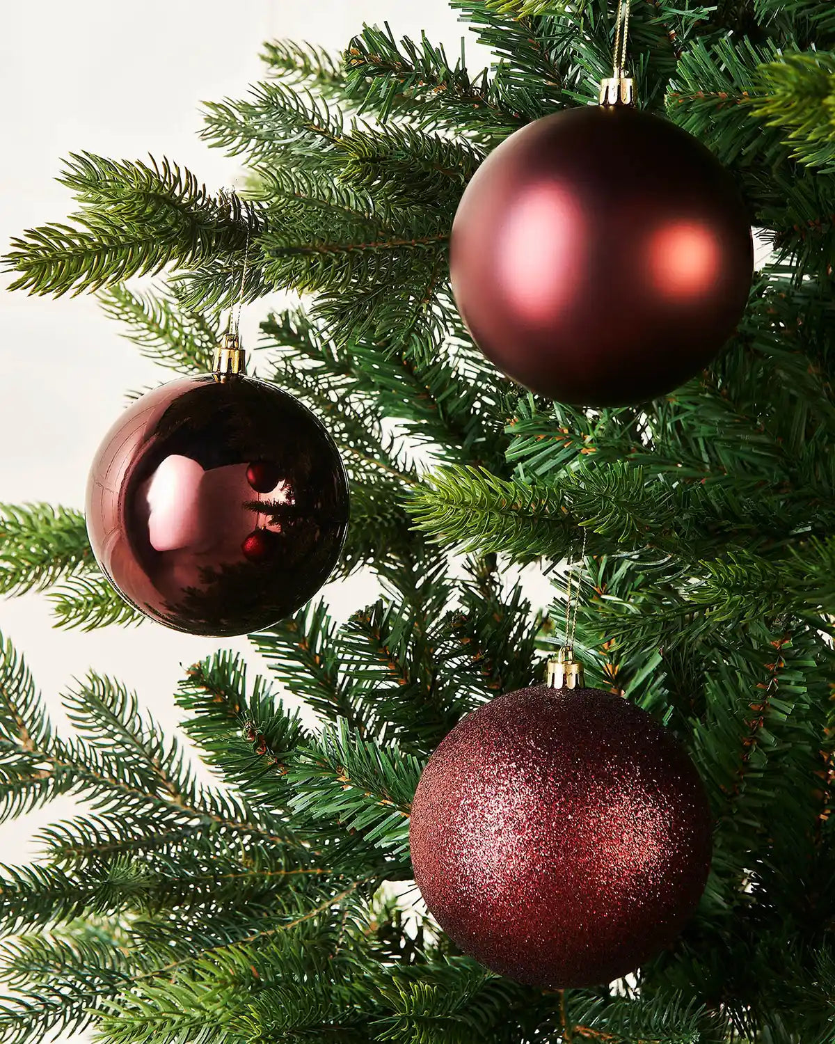 Three burgundy Christmas ornaments on a green tree with a white wall
