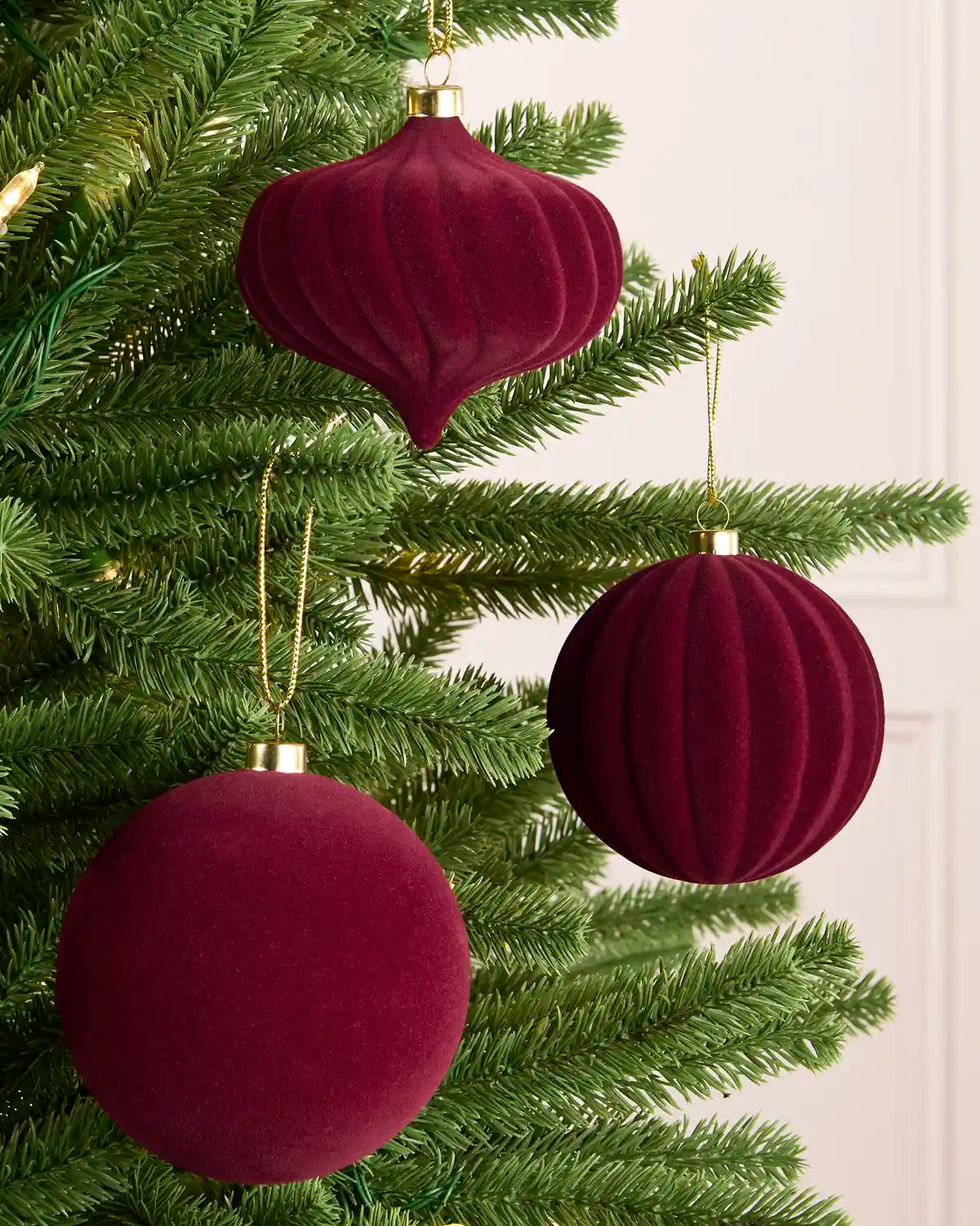 Three burgundy velvet Christmas tree ornaments hanging on a green pine tree, including two ribbed round baubles and one ribbed teardrop ornament with gold hanging loops.