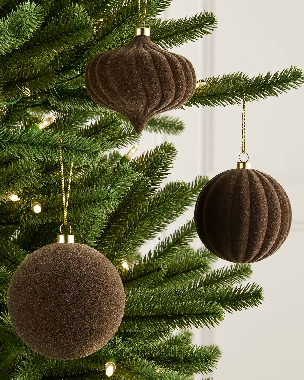 Three dark brown textured Christmas tree ornaments, including two ribbed balls and one ribbed teardrop, hanging on a green pine tree with warm string lights.