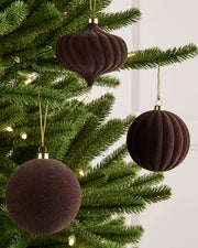 Three espresso coloured velvet Christmas tree ornaments, including two ribbed teardrop shapes and one smooth round ball, hanging from a green Christmas tree with warm white string lights.