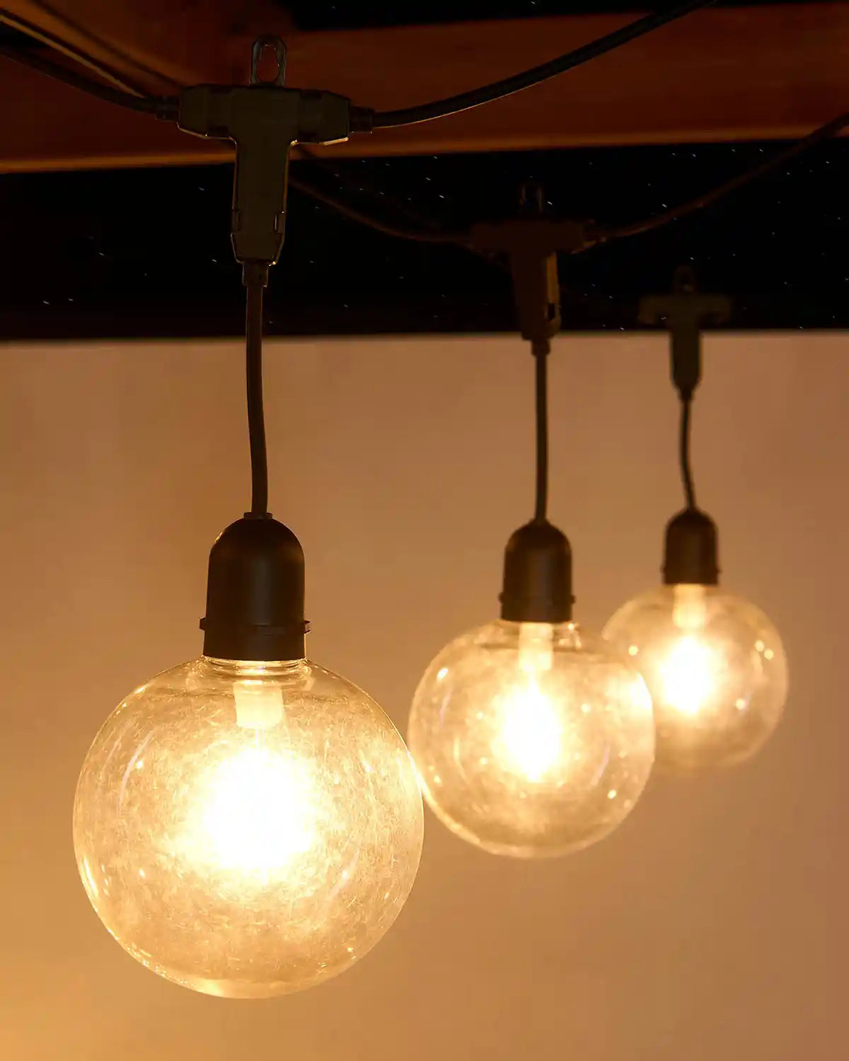 Three illuminated light bulbs hanging from a wooden beam on a ceiling fixture against a neutral background