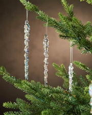 Three iridescent crystal icicle ornaments hanging from a Christmas tree branch against a brown background  