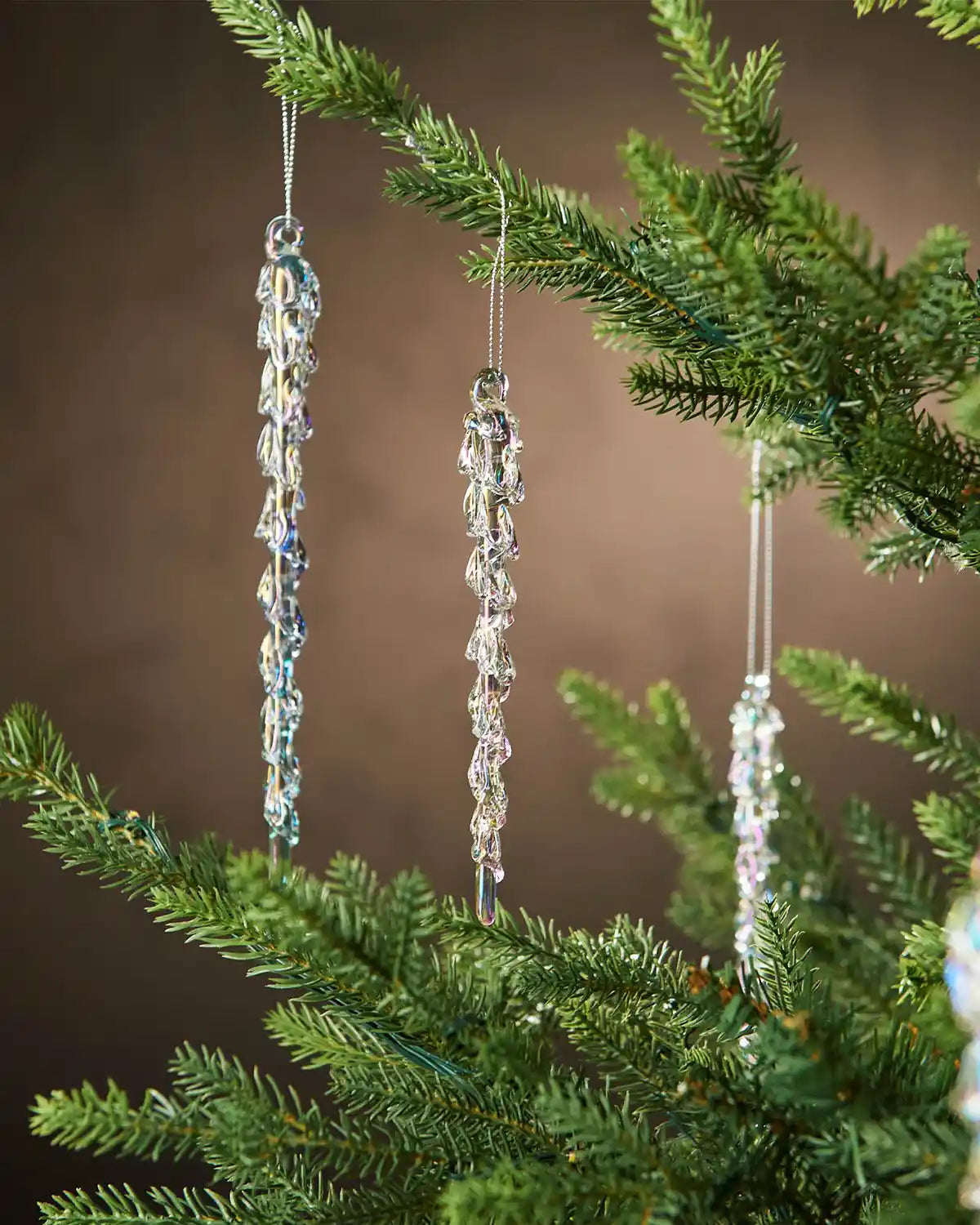 Three iridescent crystal icicle ornaments hanging from a Christmas tree branch against a brown background  