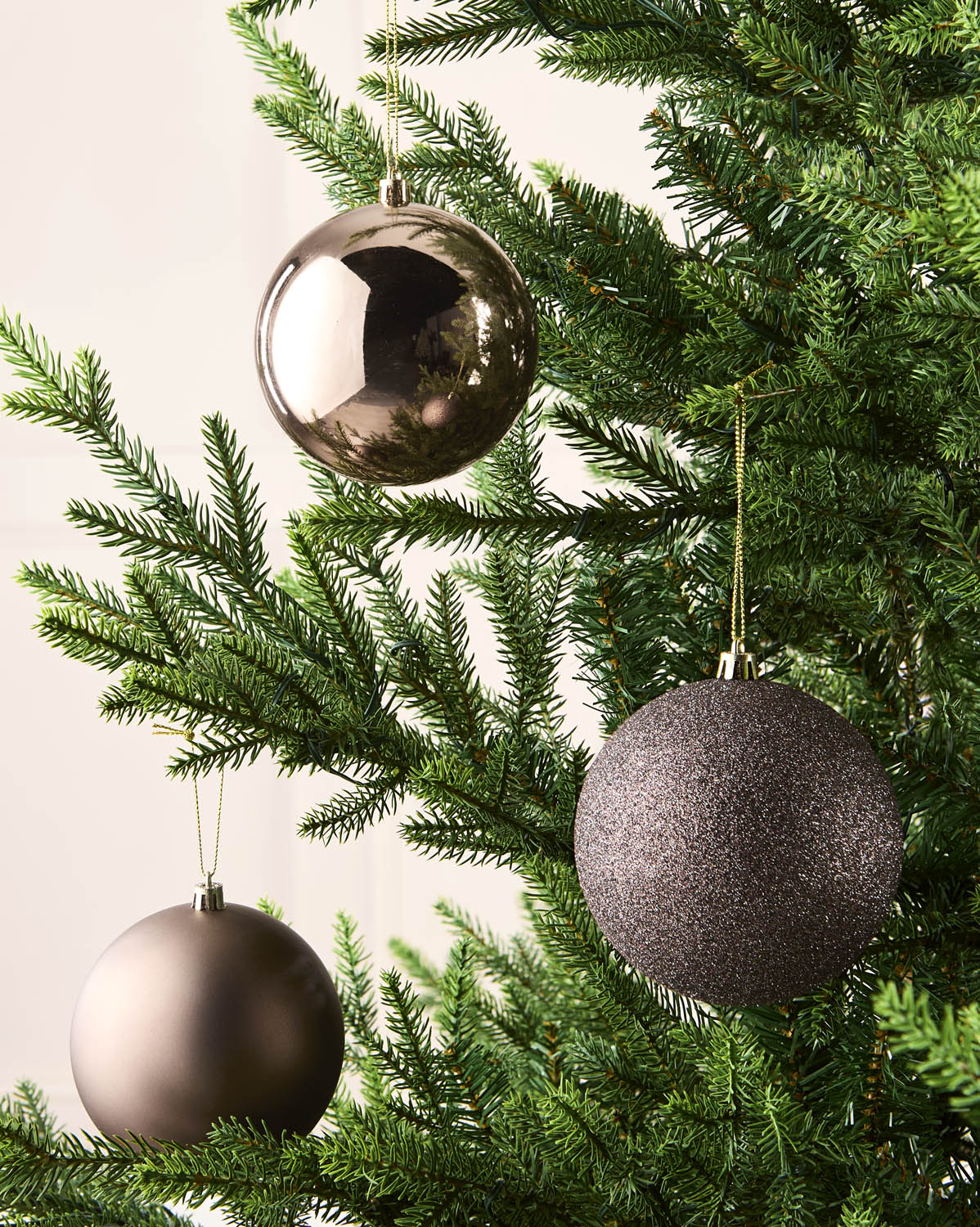 Three metallic Christmas tree ornaments in Mocha colour and gold finishes hanging on green pine branches against a light neutral background.