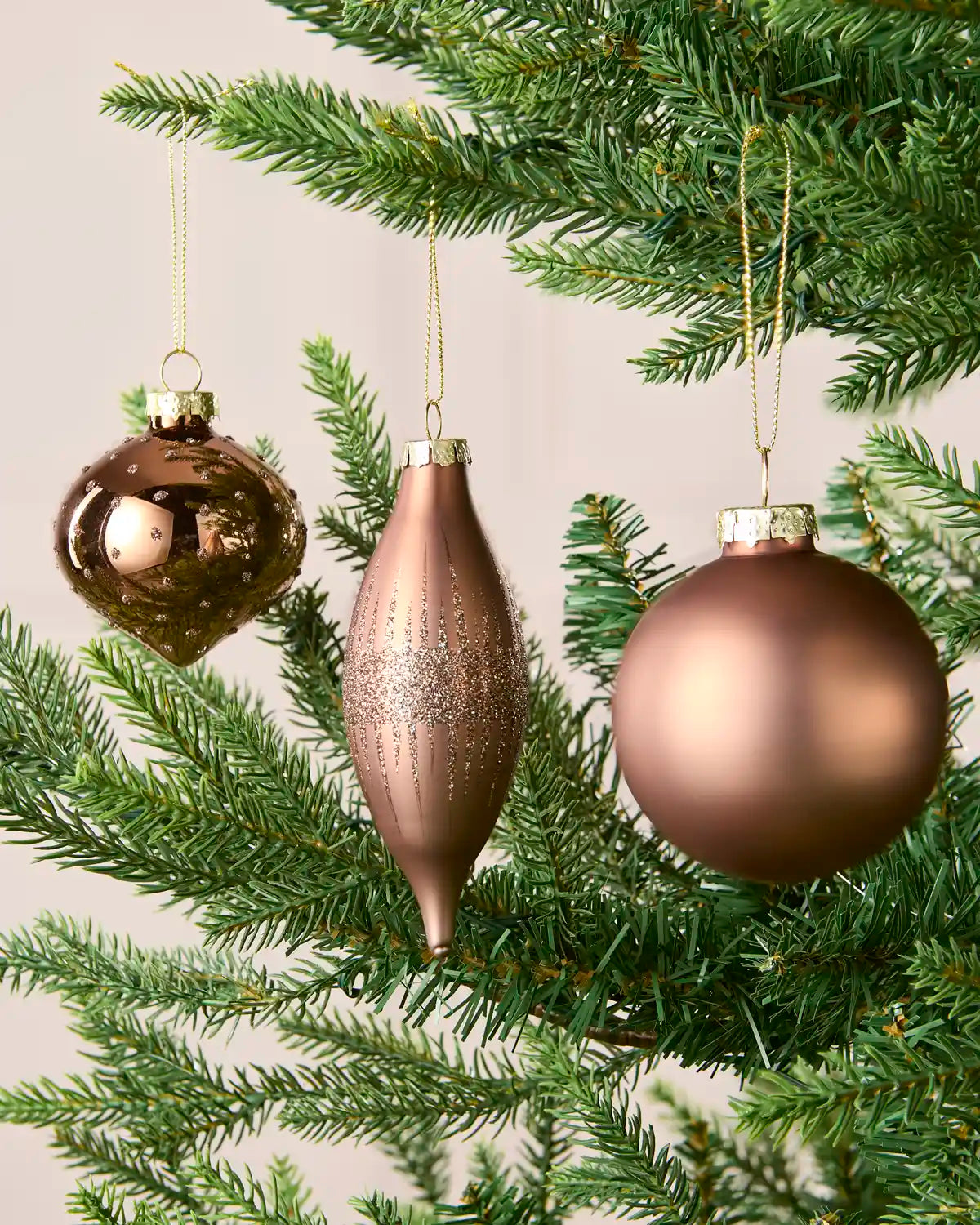 Three metallic Christmas tree ornaments in Mocha tones hanging from green pine branches against a soft beige background.