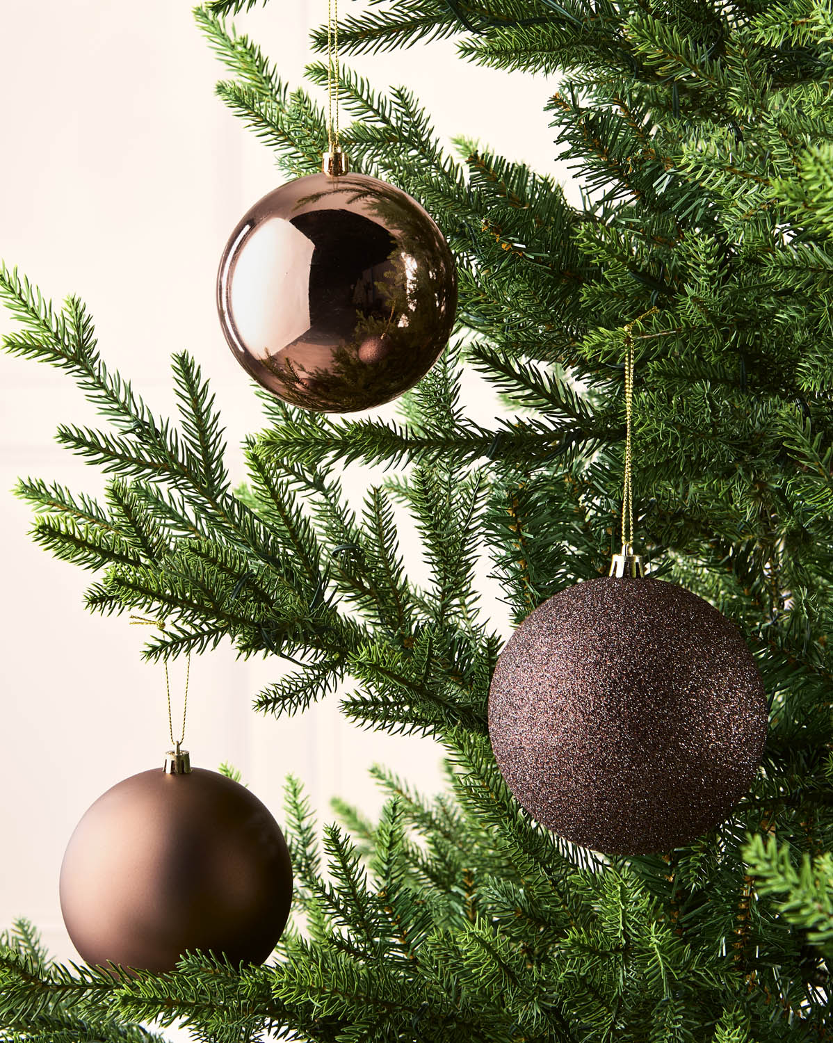 Three metallic Christmas tree ornaments in espresso finishes hanging from the green branches of a Christmas tree.