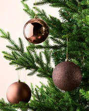 Three metallic Christmas tree ornaments in espresso finishes hanging from the green branches of a Christmas tree.