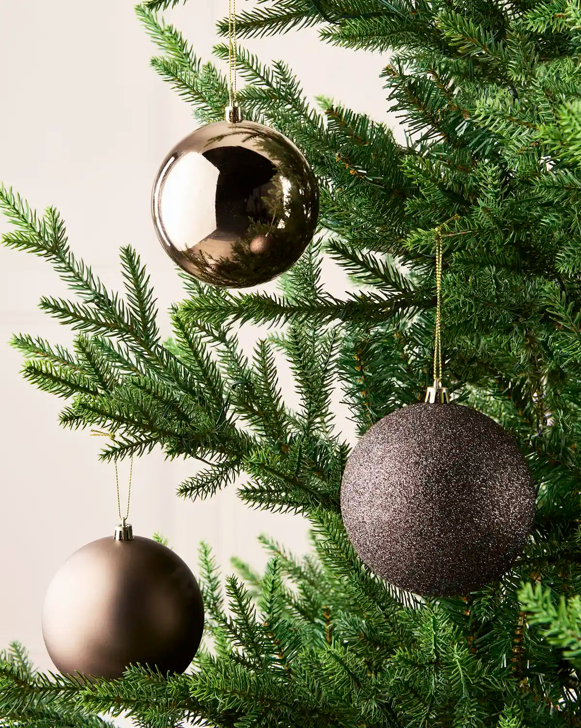 Three metallic Espresso, Christmas tree Baubles, textured and satin bronze finish hanging on a green pine tree against a light neutral background.