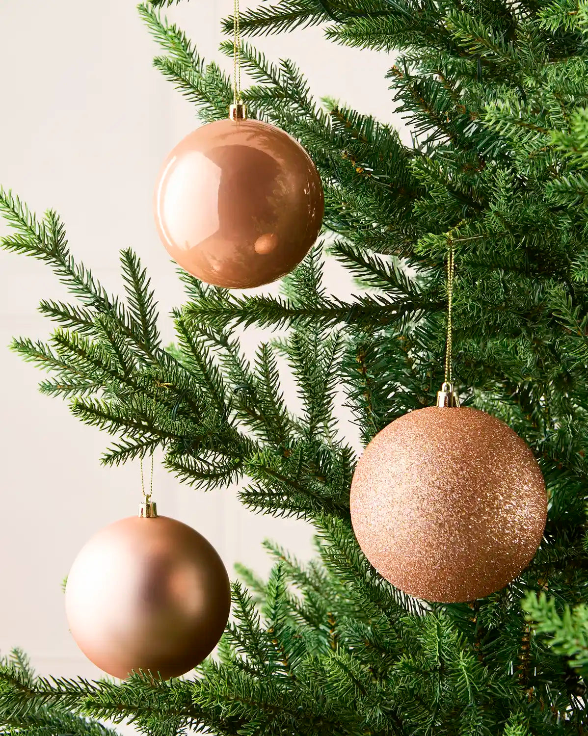 Three metallic rose gold Christmas tree ornaments hanging on a green artificial Christmas tree against a light neutral background.