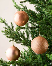 Three metallic rose gold Christmas tree ornaments hanging on a green artificial Christmas tree against a light neutral background.