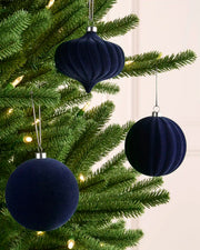 Three navy blue velvet Christmas ornaments, including two ribbed spheres and one ribbed teardrop shape, hanging on a green Christmas tree with warm string lights.