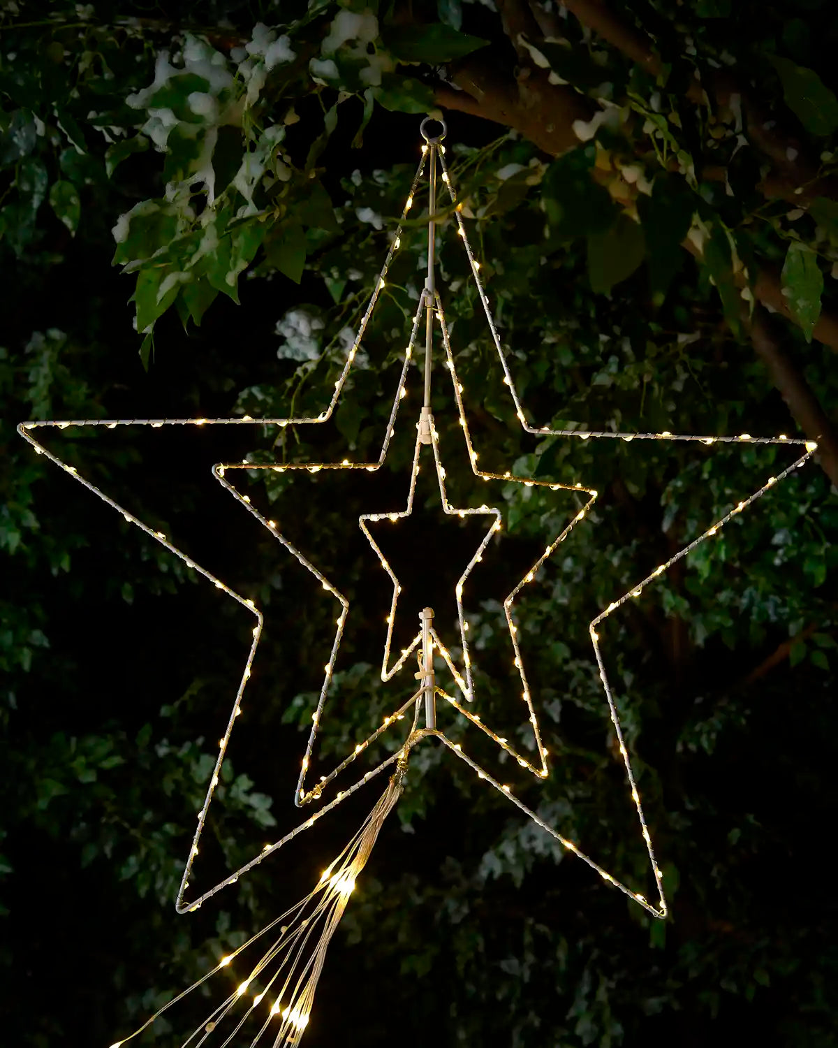 Three nested star-shaped LED light decorations glowing with warm white light, hanging from tree branches outdoors at night.
