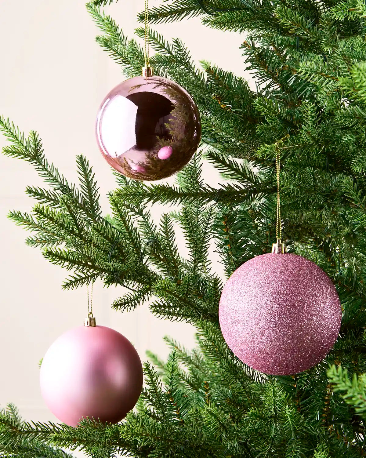 Three pink baubles with gold string hanging on a Christmas tree against a cream background