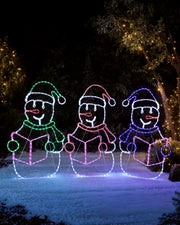 Three pre-lit carolling snowmen rope light silhouettes glowing in colorful lights outdoors on snow-covered ground