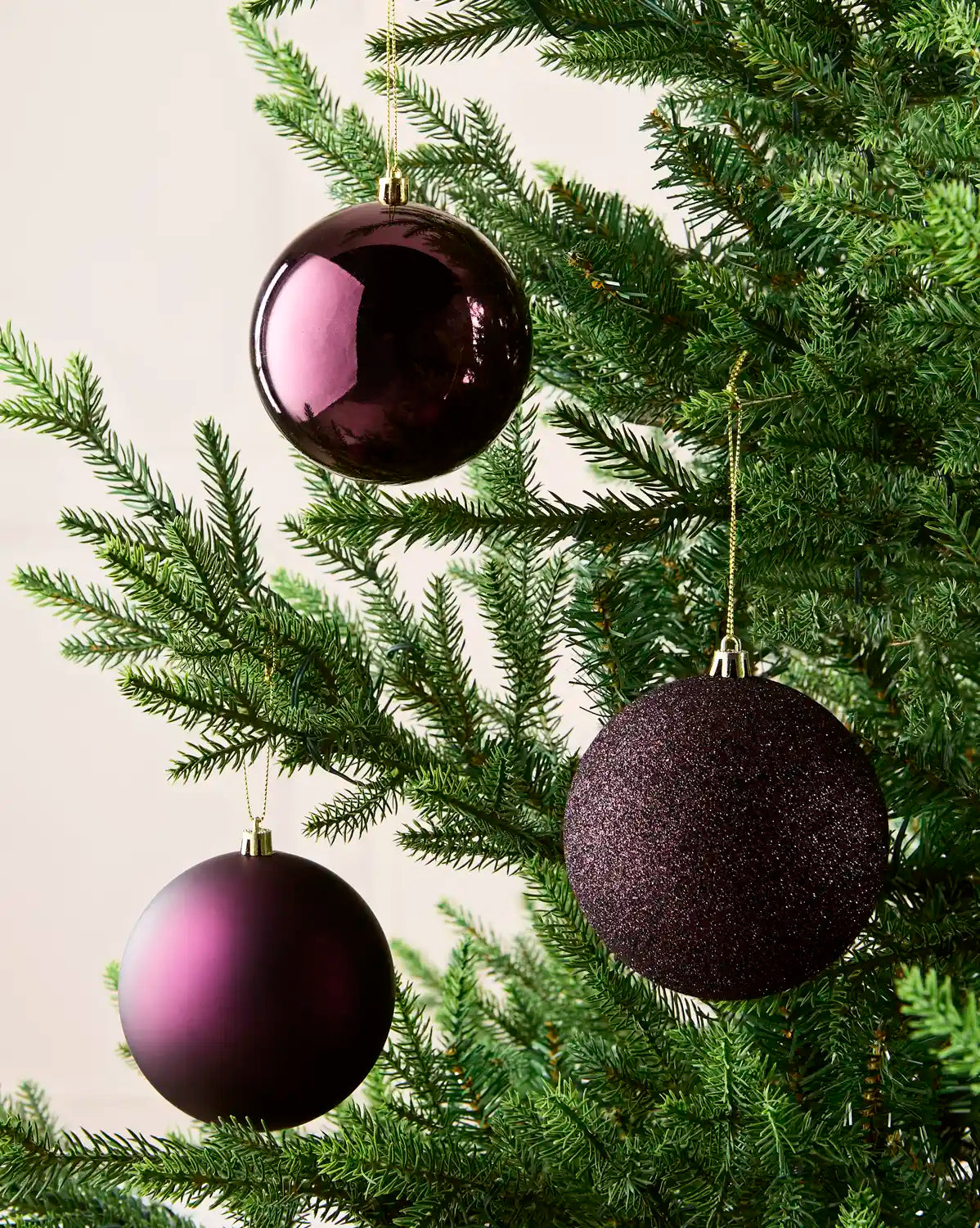 Three purple Christmas Baubles with gold string hanging on a Christmas tree against a beige background