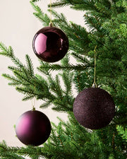 Three purple Christmas Baubles with gold string hanging on a Christmas tree against a beige background