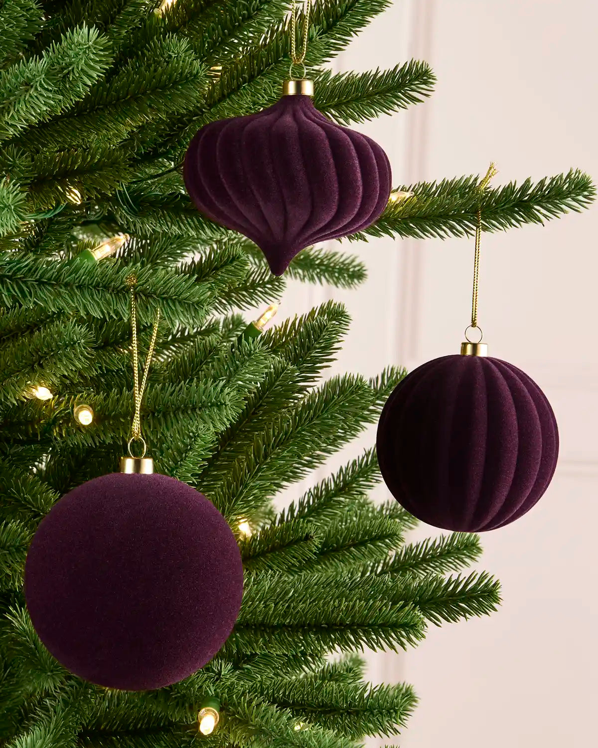 Three purple velvet Christmas ornaments, including two ribbed round baubles and one ribbed teardrop ornament, hanging on a green Christmas tree with warm string lights.