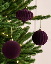 Three purple velvet Christmas ornaments, including two ribbed round baubles and one ribbed teardrop ornament, hanging on a green Christmas tree with warm string lights.
