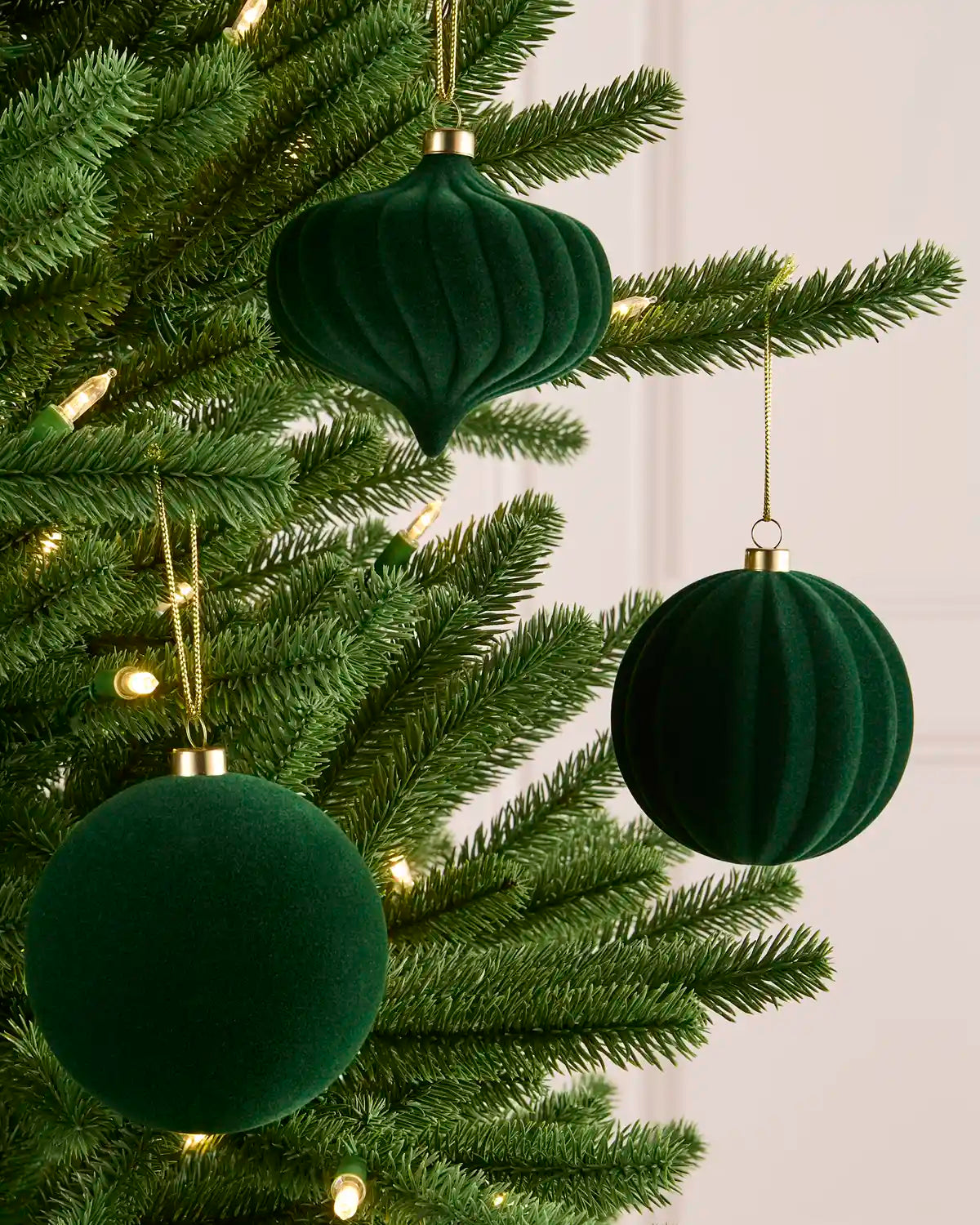 Three sage green velvet Christmas tree ornaments hanging on a green pine tree with warm white string lights against a soft off-white panneled wall.