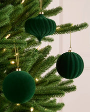 Three sage green velvet Christmas tree ornaments hanging on a green pine tree with warm white string lights against a soft off-white panneled wall.